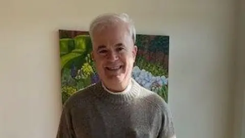 Police Scotland A man with short white hair smiles at the camera. He is wearing a grey jumper and stands in front of a painting hanging on a wall