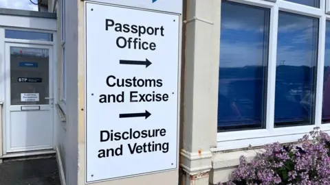 À sign on a wall says: Passport Office, Customs and Excise, Disclosure and Vetting with arrows pointing right.