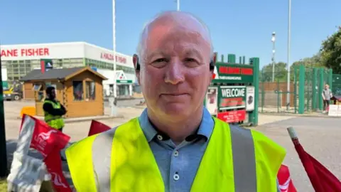 A man looking at the camera is wearing a yellow high-vis vest and a blue polo top. 