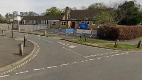 A single story pale brick building with a playground and parking spaces to the foreground, surrounded by wire fencing. A blue sign stands inside the school grounds, situated on a corner of a road.
