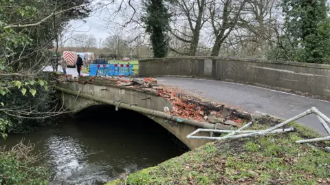A general view picture of the bridge, which has seen its side closest to the camera totally flattened, with metal railings bent out of place and bricks scattered nearby.