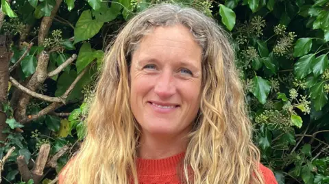 A woman with long blond hair in an orange jumper standing in front of a hedge