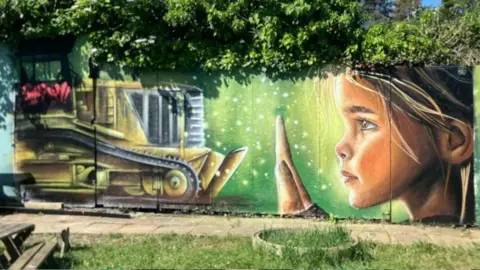 SHARIQUA AHMED/BBC Mural of a girl holding up a hand to a yellow bulldozer- with a green background painted on a wall lined with trees.