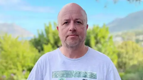Man with a shaved head and light stubble stands outdoors in front of blurred greenery and distant mountains, looking directly at the camera with a neutral expression. He is wearing a light grey T-shirt, and the scene is brightly lit with natural daylight.