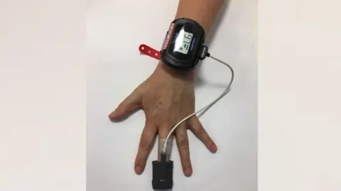 A hand is in the middle and is on a table. A person is wearing a black monitor device on their wrist which has a wire linked to a finger monitor.