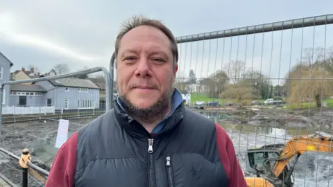 Shaun Whitmore/BBC Jonathan Little wearing a black gilet and maroon jumper. He has short brown hair and a beard and is smiling while stood in front of the digger, which is stuck in mud.