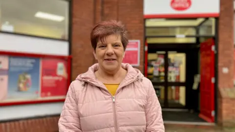 Jane Ellison stands outside the entrance of the Redditch branch, wearing a pink puffer jacket and yellow top.