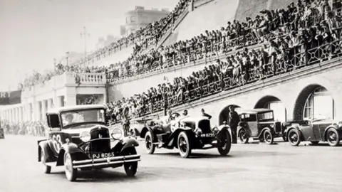 The Regency Society An old black and white photograph of two cars racing on a road, with hundreds of spectators looking on. 