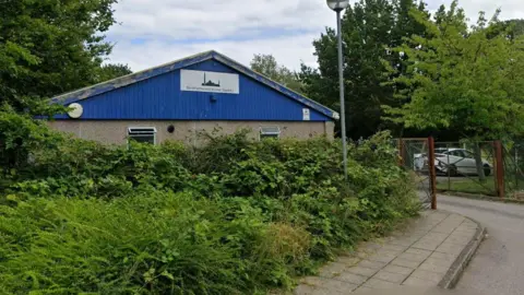 A single-storey building with blue cladding stands behind dense greenery and shrubs beside a narrow road. A sign on the front of the building reads “Borehamwood Islamic Society”. A lamppost stands in the foreground, while a metal gate to the right leads to a parking area where a white car is visible. Trees surround the site under a cloudy sky.