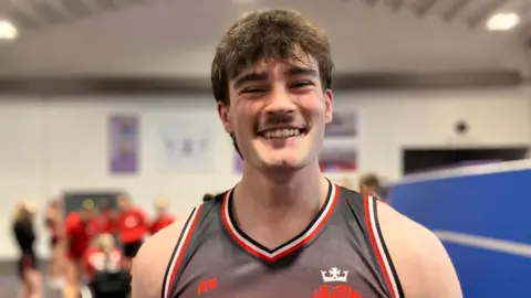 Harrison wears a sleevless training top with red and black on it. He has brown hair and a moustache