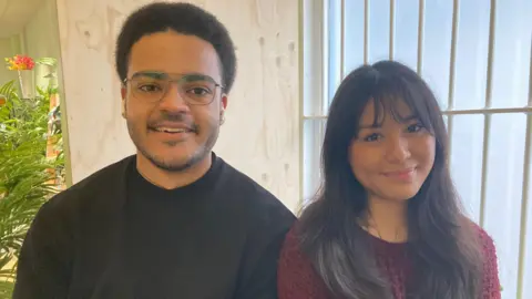 Year three students Ethan Jessamy is wearing a black sweatshirt and is sitting next to Jenny Gauchan who is wearing a burgundy jumper. They are standing in front of a cream-coloured wall with a window that has bars on it. A floral display can be seen at the left edge of the shot. 