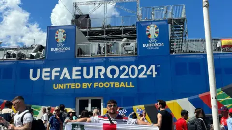 Casey Bishop Casey Bishop stood outside a book stadium with the words 'UEFA Euro 2024, united by football' written in white letters. 
