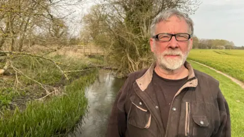 Greg Knowlson, who lives next to the treatment works at South Moreton in Oxfordshire, poses for a photo near the brook. He is wearing a brown jacket and dark-rimmed glasses. It is cloudy.