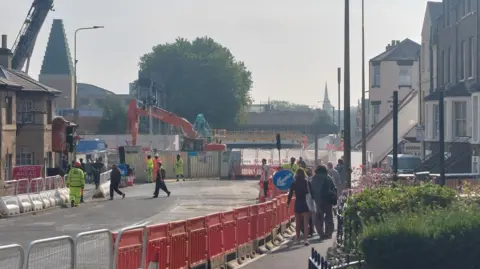 People seen walking near the Westgate Hotel on Botley Road on the approach to the centre of Oxford. There are rows of orange barriers placed along the road ad several workers in yellow high vis uniforms are around. It is a foggy day.