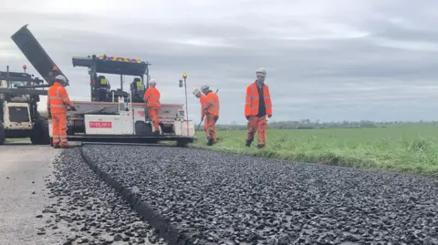 Oxfordshire County Council Highway maintenance workers use a paving machine and tipper truck to resurface a road. Fresh asphalt is being laid on a damp roadway bordered by trees, with safety lights visible on the machinery.