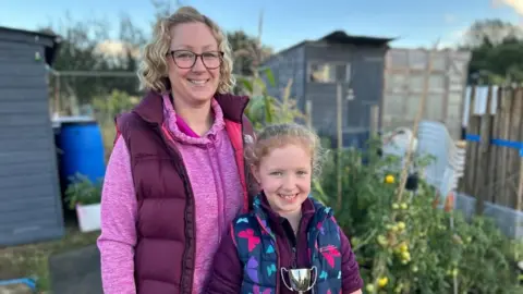 Kate Bradbrook Keeley standing wearing pink jumper, purple gilet, with short blonde her and glasses, with her daughter Penny