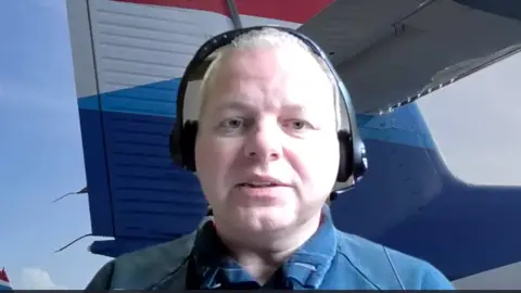 Jonathan Hinkles is talking and wearing headphones. He is sitting in front of a photo of an airplane's tail fin.