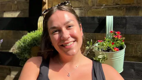 Harriet Heywood/BBC Ms Marsh is smiling at the camera while wearing a black top, earrings and silver necklace. She has long brown hair and sunglasses on top of her head. She sat outside in front of a brown fence and the sun is shining on her face. Behind here are some hanging baskets with flowers planted inside.