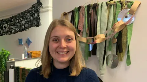 A woman with blond shoulder length hair is wearing a navy blue jumper and smiling. She is stood in front of a wall which has paperchains and fabric decorations