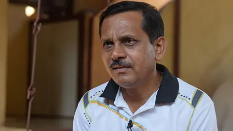 Head and shoulder shot of Daji Rajguru - he has short dark hair and a moustache. He is wearing a white sports shirt with a dark blue collar and some blue and yellow markings.