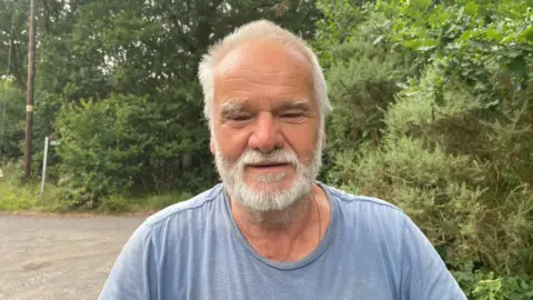 Lawrence Porter, a resident of Woodhall Spa, talking about the closure of the car park at Ostler's Plantation. He has a grey beard and hair and is wearing a blue t-shirt.