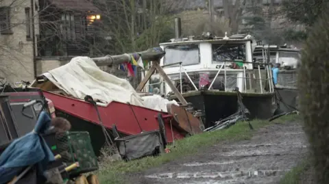 The picture is taken from the path next to the canal where boats are lined up. Some looked damaged or in poor condition with bikes laid on the ground next to the boats and a a small bike trailer upside down. 