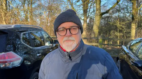 Nathan Turvey/BBC A picture of James Jackson, wearing glasses, a blue hat and coat, with cars either side and trees in the background.