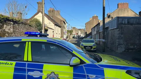 Cillian Sherlock/PA Wire Two garda (Irish police) cars parked at the cordon in New Ross.  The narrow street is lined with terraced houses of different heights.   A wall in the foreground is covered with overgrown plants.