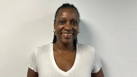 A woman with black hair and wearing a white T-shirt and a gold necklace smiling at the camera against a plain white background.