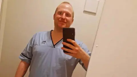 Sheena Millar A white blonde man in his 30s is pictured taking a selfie in the mirror. He is smiling and wearing a light-blue NHS-branded uniform.