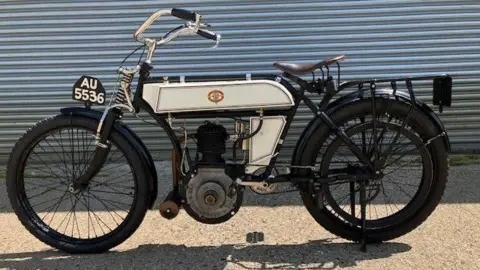 A black and white 1904 Riley motorcycle, parked in front of a corrugated silver shutter.