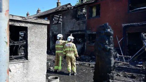 Pacemaker Two firefighters in yellow uniforms and white helmets in front of the burnt out red and white houses. 