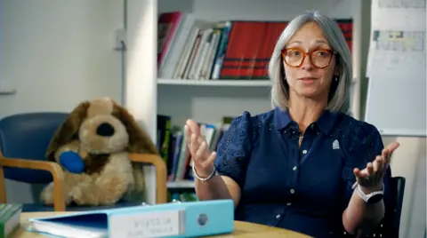 A woman with shoulder length grey hair and glasses in a dark blue blouse. She sits in an office with a teddy dog on a chair behind her.