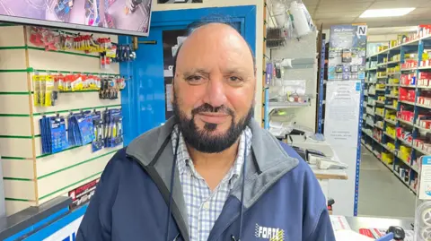 A man with a dark beard wearing a blue jacket stands in the shop he owns. He is stood in front of the counter, behind it are racks of shelving.