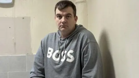 BBC Andrew Gibney has short black hair and is wearing a grey hoodie with BOSS white letters logo.