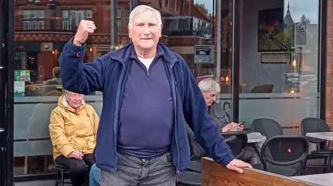 Rob Cocker A man with short white hair and a moustache, wearing a blue jacket, jumper and jeans, raises his right fist in the air in celebration while standing outside a pub.