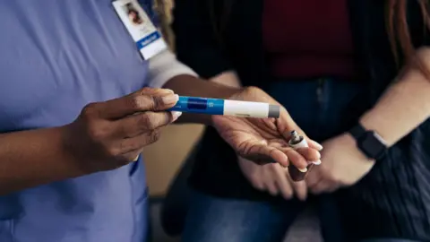 Getty Images Close-up of nurse's hands holding a medical pen-like device for self-injection of weight loss drugs. In the background is a male patient sitting on an exam bed. 