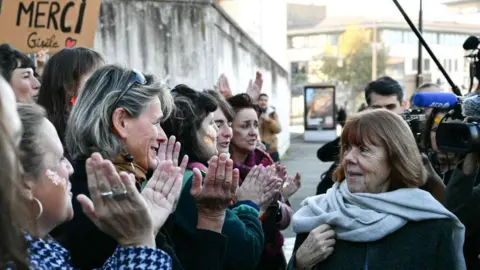 CHRISTOPHE SIMON/AFP via Getty Images Gisèle, smiling slightly, walks past a group of women who are applauding her. In the background someone holds up a sign that says "merci Gisèle".