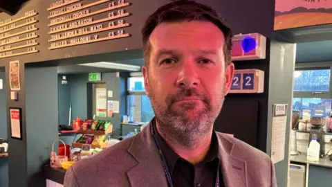 Dave McPartlin has dark hair and a greying bard. He wears a dark blue polo shirt and brown jacket.