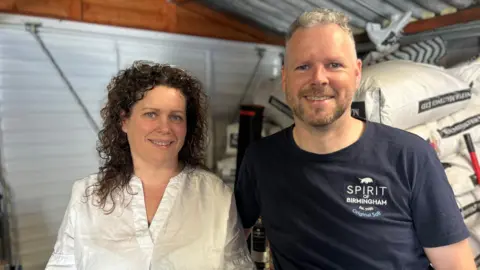 A woman with brown curly hair is looking at the camera smiling. She is wearing a white v-neck shirt. Next to her is a man with short grey hair who is also smiling at the camera. He is wearing a blue t-shirt with "Spirit of Birmingham" on it. Behind them is a white garage door and white bags of barley. 