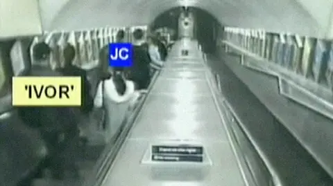 CPS - Crown Prosecution Service CCTV image from Stockwell tube station showing Jean Charles de Menezes, who is marked "JC", on an escalator, being followed by a surveillance officer, who is marked "IVOR". There are framed images on the sides of the up and down escalators.