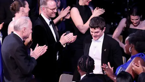 Getty Images Robert Aramayo walks to the stage as he wins the leading actor award for 'I Swear' during the Bafta film awards. He is surrounded by people applauding him.