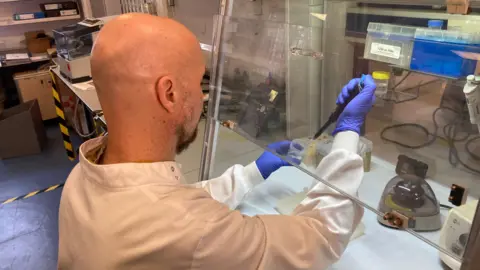A man in a white lab coat works in a laboratory. He is moving liquid from a large pipette into a container. 