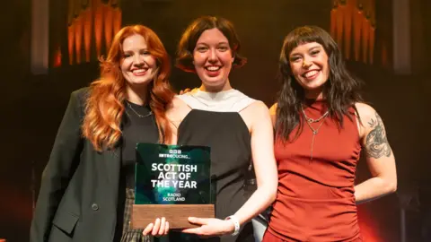 Sean Purser Three smiling women stand on a warmly lit stage in front of organ pipes, with the woman in the centre holding a glass and wooden trophy that reads “BBC Introducing… Scottish Act of the Year – Radio Scotland”. The woman on the left has long red hair and wears a dark blazer and skirt, while the woman on the right has dark shoulder-length hair, a sleeveless red outfit, and visible arm tattoos.