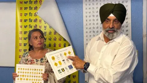 Gagandip Singh Um cara idoso indica um turbante preto e uma camisa branca com símbolos em uma folha de papel em uma folha de papel. Uma mulher idosa com roupas de flores ao lado dela, contém uma folha semelhante com vários símbolos. Eles estão na frente de dois pôsteres com letras de Punjabi