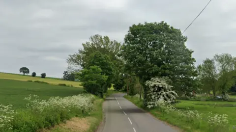 A general view of the B966 at Cairnton of Balbengo. The road cuts through the middle of the image and is grey with white markings on it. There is grass and fields on either side and in the middle of the image, trees going overhead. They sky is grey.