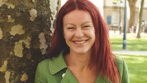 @JonCraig photos Christine Townsend with red hair, leaning against a tree and smiling at the camera