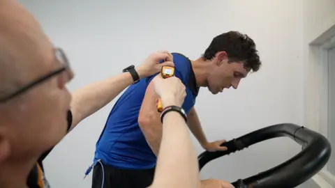 A man in a blue shirt on a treadmill. Another man is holding a probe up to his right shoulder.
