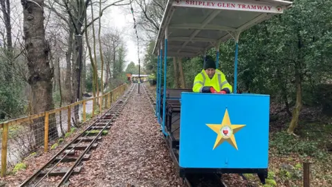 A man in a fluorescent yellow jacket and black hat riding in a blue tram carriage