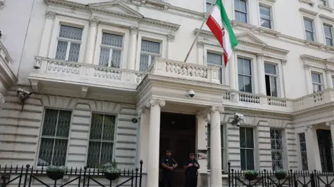 Two police officers stand outside a white Iranian Embassy building flying the Iranian flag, with barred windows and flower baskets on the fence.